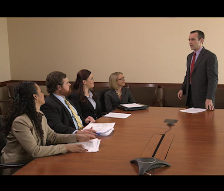 Video still image of the introductory video of John Moore standing in front of his colleagues at a conference table.